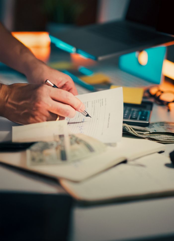 brand-02 A hand signing financial documents on a desk with cash and a calculator, signifying finance management.